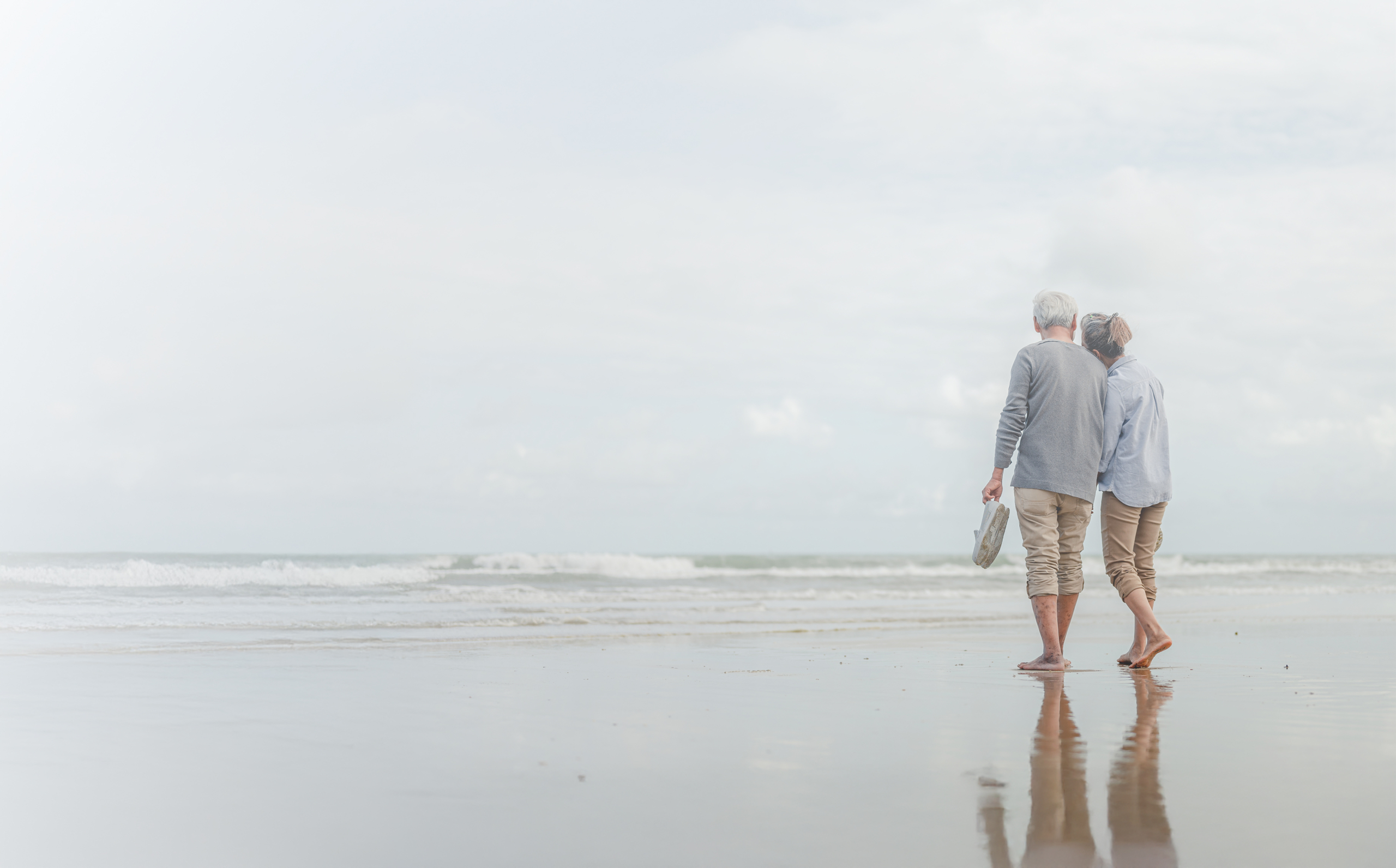 An older couple walking together barefoot on a peaceful beach, facing away from the camera, with gentle ocean waves and a calm sky in the background.